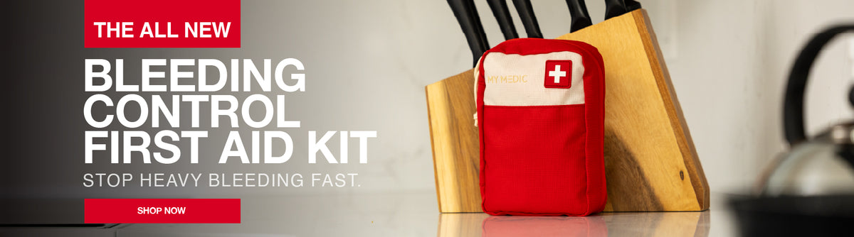 A red first aid kit pouch with a white cross on the front stands on a countertop beside a wooden knife block. Large promotional text on the left reads 'THE ALL NEW BLEEDING CONTROL FIRST AID KIT' with a red 'SHOP NOW' button.