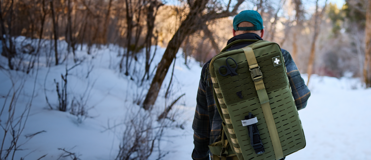 A person in a snowy outdoor setting viewed from behind, wearing a green tactical backpack with a white cross symbol on the back.