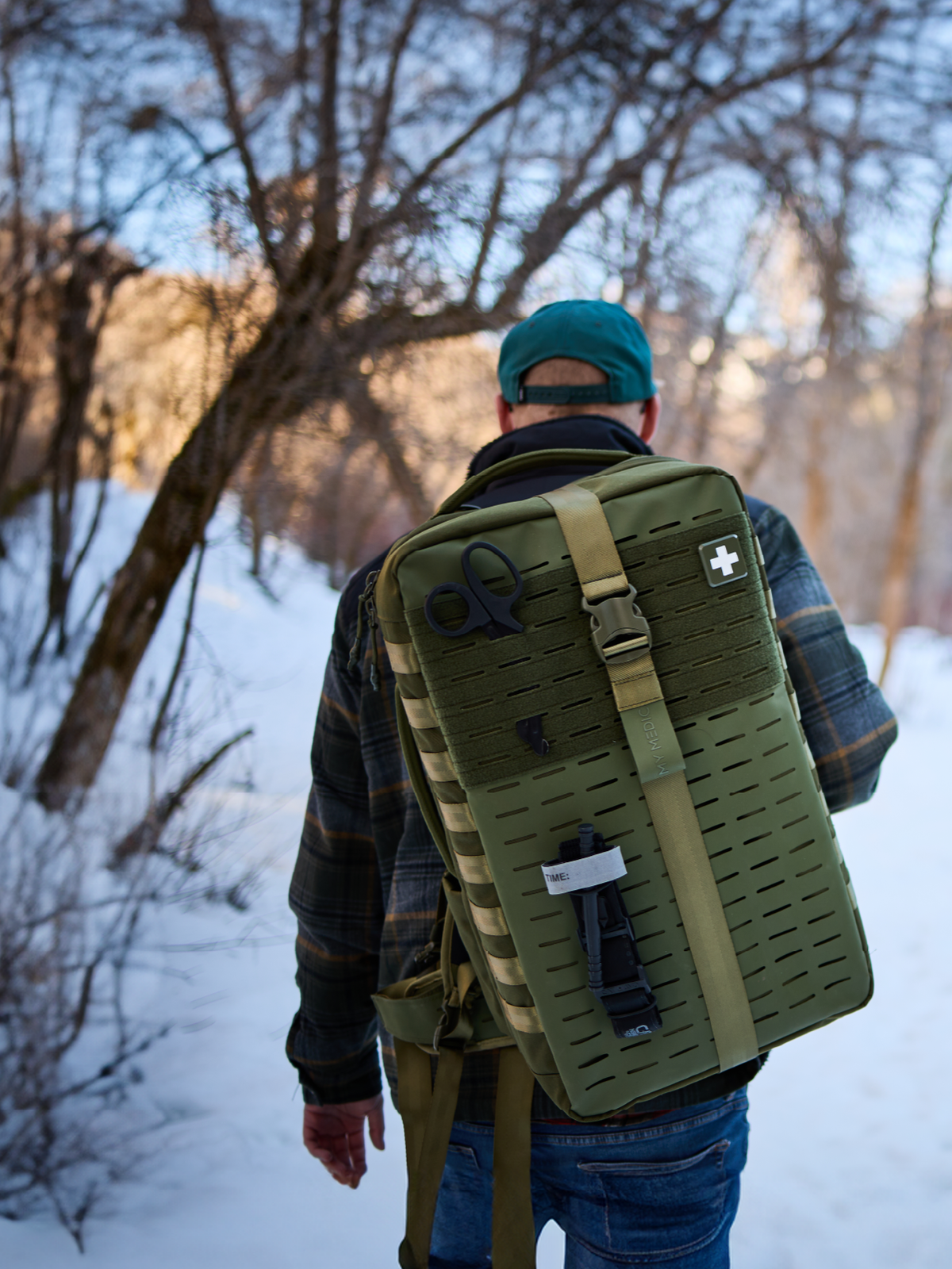 A person viewed from behind walking through a snowy outdoor setting, wearing a large olive green tactical backpack with a padded rectangular panel, straps, and a white cross patch.