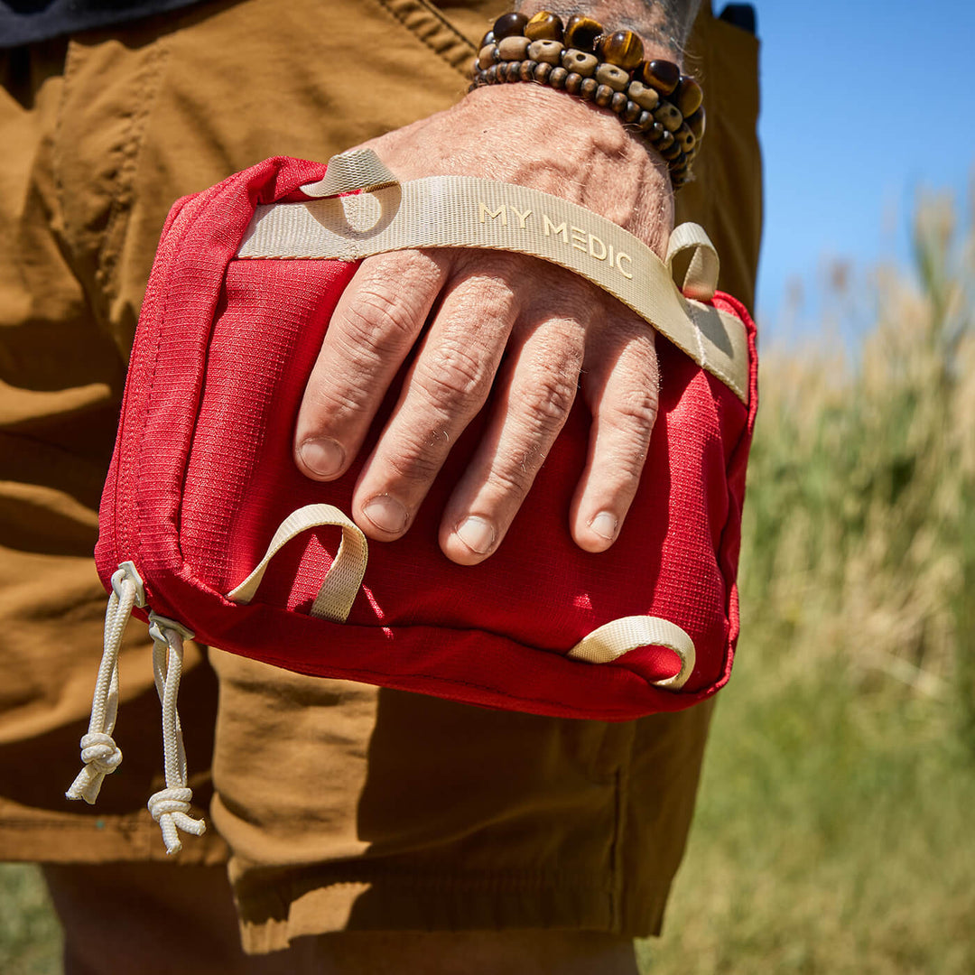 A close-up of a person outdoors holding a red first aid pouch with a beige wrist strap labeled 'MY MEDIC'. The person is wearing tan shorts, and the background shows blurred green grass and blue sky.