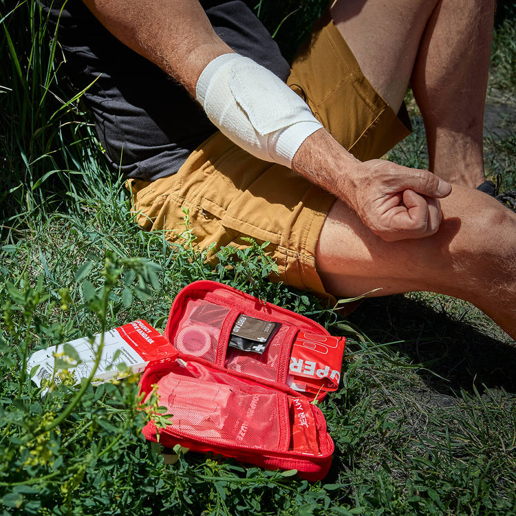 An open red first aid kit lies on grass beside a person sitting outdoors, with the person's arm wrapped in an emergency pressure bandage and shorts in tan. Visible kit contents include gloves, gauze, and other medical supplies.