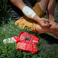 An open red first aid kit lies on grass beside a person sitting outdoors, with the person's arm wrapped in an emergency pressure bandage and shorts in tan. Visible kit contents include gloves, gauze, and other medical supplies.