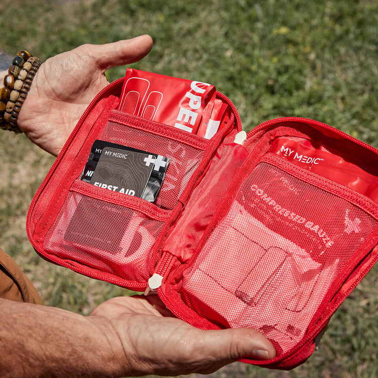 A red first aid kit pouch held open by two hands outdoors on grass. The kit contains multiple compartments with medical items, including a card labeled "First Aid & Survival Guide" and another labeled "MY MEDIC Compressed Gauze"