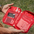A red first aid kit pouch held open by two hands outdoors on grass. The kit contains multiple compartments with medical items, including a card labeled "First Aid & Survival Guide" and another labeled "MY MEDIC Compressed Gauze"