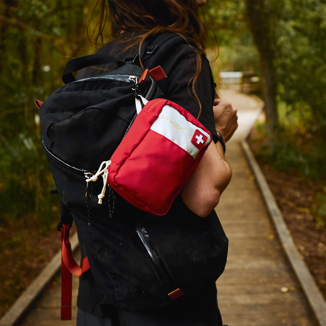 A person wearing a black backpack on a wooded path, carrying a red first aid pouch on their shoulder. The pouch has a white top section with a white Swiss cross and the brand name 'My Medic' printed on it.