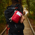A person wearing a black backpack on a wooded path, carrying a red first aid pouch on their shoulder. The pouch has a white top section with a white Swiss cross and the brand name 'My Medic' printed on it.