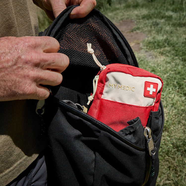 A close-up of a black backpack being worn outdoors, with a person's hands holding it open to show a red and off-white MY MEDIC first aid pouch inside a side pocket.