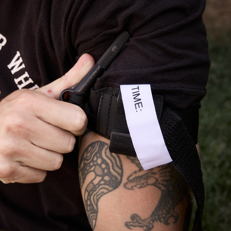 A close-up of a person's tattooed arm wearing a black tactical-style wrist strap or holster, with a white paper tag printed with 'TIME:' attached. A hand is adjusting the black strap over a black T-shirt.