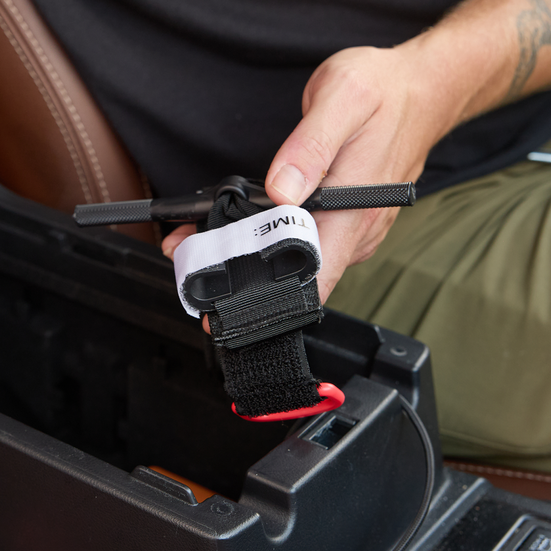 A close-up of a person's hand holding a black car center console armrest organizer insert with a white and black strap and a red carabiner clip, shown installed in a black console compartment.