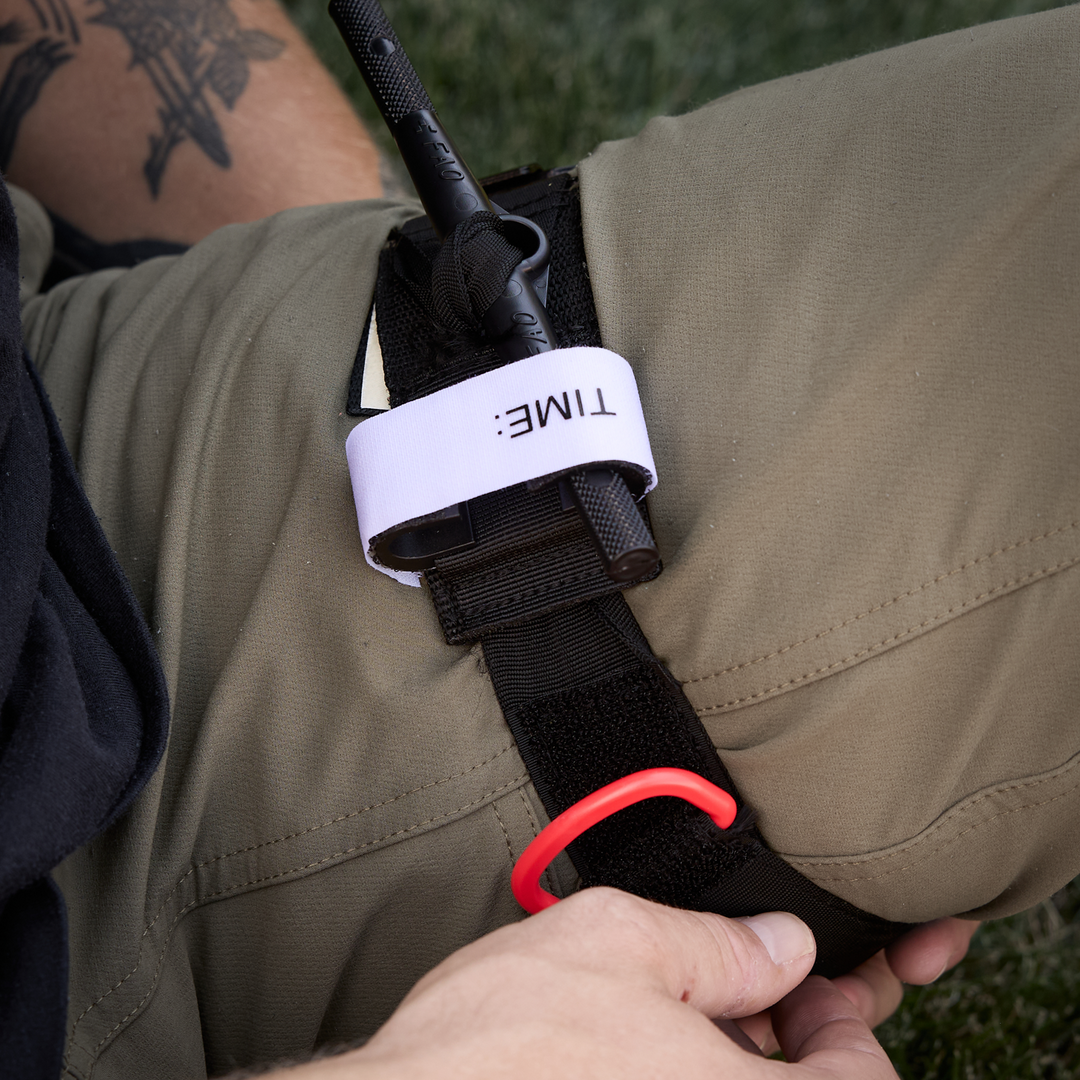 Close-up of a person wearing olive green pants adjusting a black windlass tourniquet. 